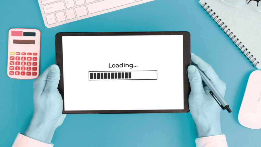 Hands holding a tablet displaying a loading screen with a progress bar. Surrounding items on the blue desk include a keyboard, calculator, notepad, glasses, and a white mouse. The hands are typing or using a stylus on the tablet, perhaps tweaking some WordPress plugins.