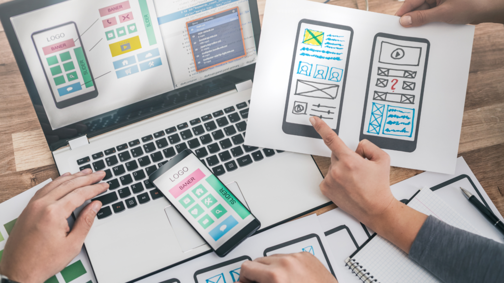 A team in Cameroon collaborates on mobile app design at a web design agency, with hands pointing at paper wireframes displaying app layouts. A laptop shows additional wireframes, a smartphone sits on the keyboard, and a notepad with sketches and notes is on the table.