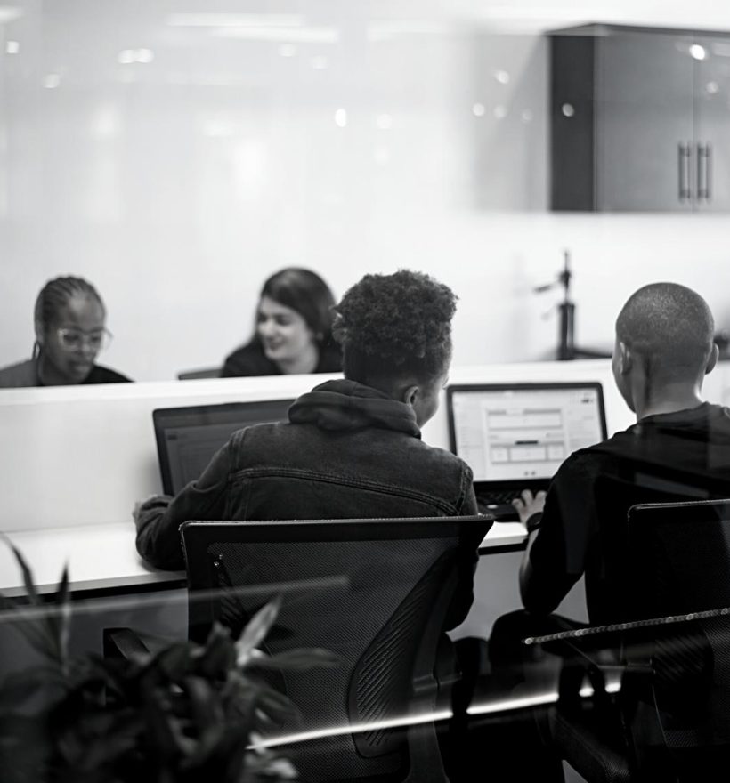 Black and white image of four people sitting at a table in a modern office space. Two individuals in the foreground work on laptops, likely focusing on weblim projects, while two others are seen in the background having a conversation. The room has a clean and minimalist design.