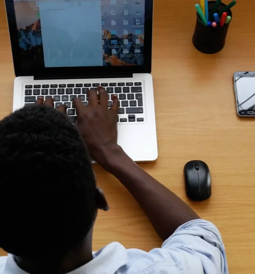 A person sits at a wooden desk, typing on a laptop. On the desk are a smartphone, a wireless mouse, a pot of colorful pens, and some papers. The scene is viewed from above, showcasing the person's hands on the keyboard and part of their head and shoulders in this weblim environment.